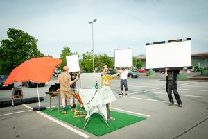 Hinter den Kulissen beim Fotoshooting "Party? Platz ist in der kleinsten Parzelle!": Zwei Männer und eine Frau halten die Reflektorpanele, in einer Parklücke liegt Rollrasen aus und Grillparty-Equipment ist aufgebaut.