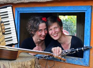 Zwei Frauen schauen durch ein blau gerahmtes Fenster, lachen und halten einen Bogen und eine Klarinette in den Händen