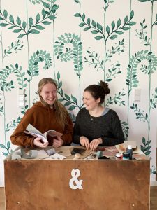 Zwei Frauen vor einer grün gemusterten Tapete, an einem Holztisch mit weißem &-Zeichen und Büchern in der Hand
