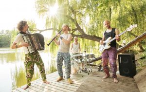 Drei Männer mit Akkordeon, Klarinette und E-Gitarre auf einem Steg an einem See, im Hintergrund Sonne, die durch eine Weide scheint